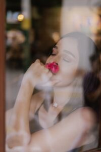 Blurred image of beautiful woman with beautiful skin, wearing sleeveless top, viewed through a glass screen or window, eyes closed and holding a rose to her face