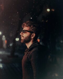 side profile of man with naturally coloured hair & beard, wearing sunglasses at dusk , in brown sweater 