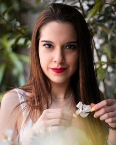 Henna hair colour is natural, photo of woman with shoulder length straight dark brown hair standing against a leafy background, holding a small white flower in her hand