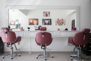 hair salon safety, why are many still using chemical hair dyes and herbal hair colours from companies whose products have killed and maimed people? ; photo of interior of hair salon, plum colour chairs facing a large mirror , reflecting poster images on a white wall