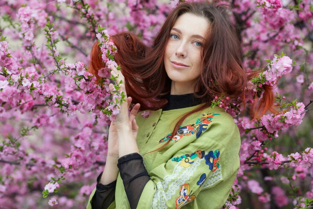 Top half of a woman with shoulder length deep auburn wavy hair, wearing a green top over a black pullover, standing in front of pink blossoms, promoting natural hair care with henna and indigo