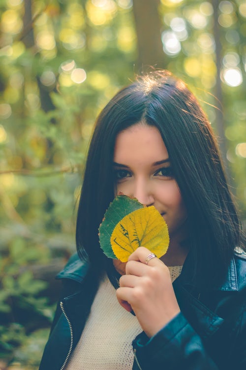 Woman with dark hair in a forest, holding up two leaves to her face, representative of how to cover grey hair naturally with henna and indigo